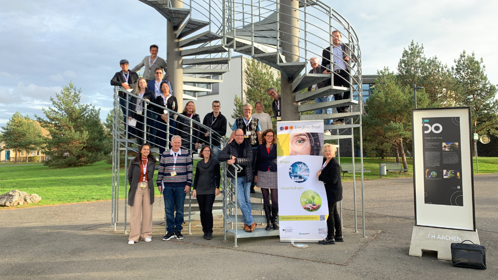 All the participants stand on and in front of a spiral staircase and smile at the photographer presenting the banner of EnergInno Brazil.